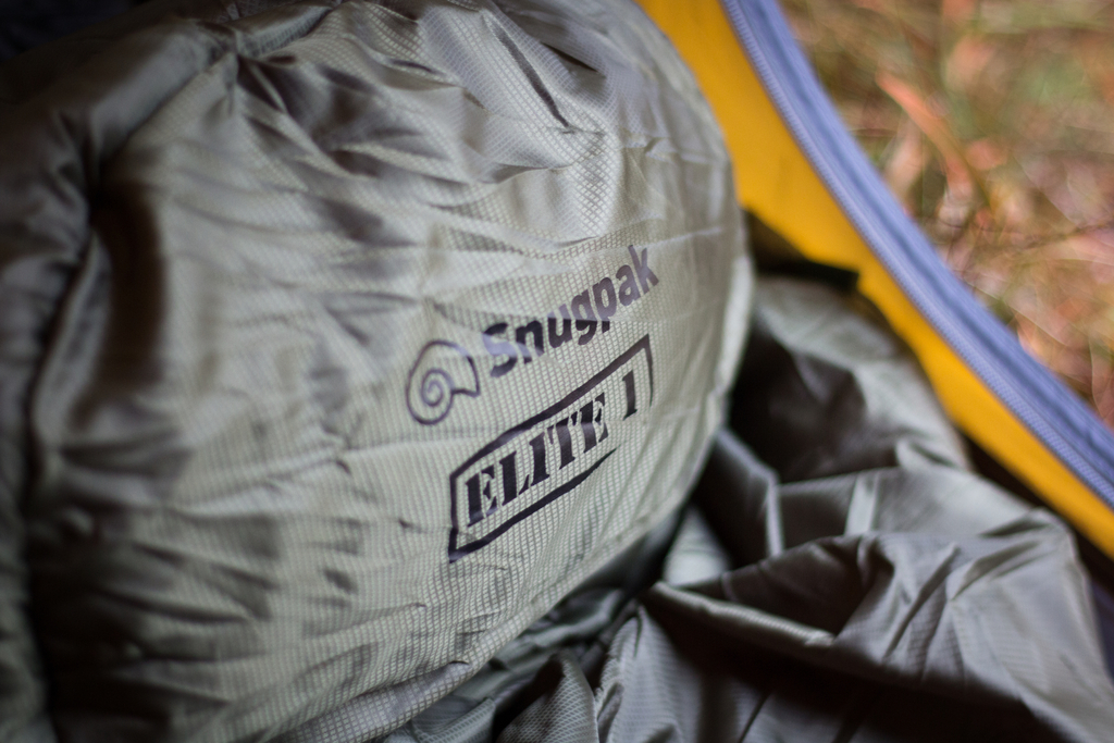 Sammenpakket Snugpak Elite 1-sovepose i sin kompressionspose ved kanten af et telt i skovbunden
