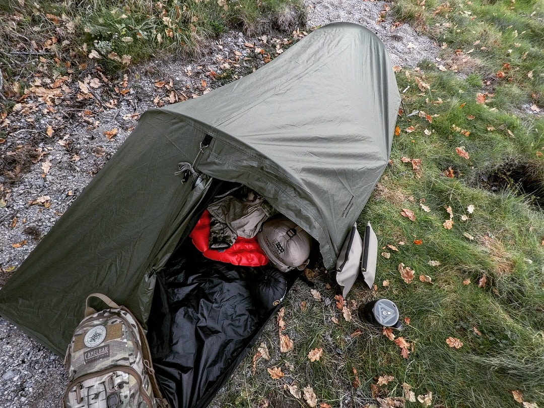 Olivengrønt enmandstelt set oppefra med åben dør, en rød og sort sovepose, en rygsæk og en kop i græsset