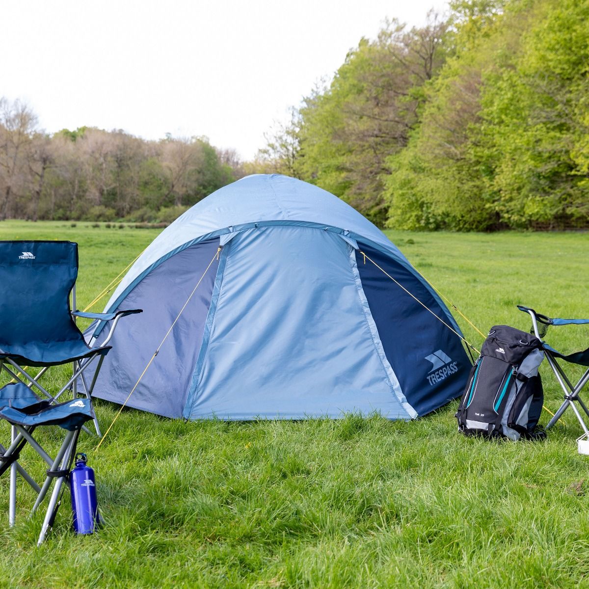 Blåt kuppeltelt slået op på en græsplæne med campingstole foran og træer bagved