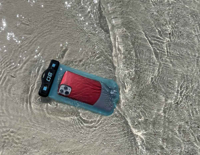 Telefon i et turkis vandtæt etui, der ligger i vandkanten på en sandstrand