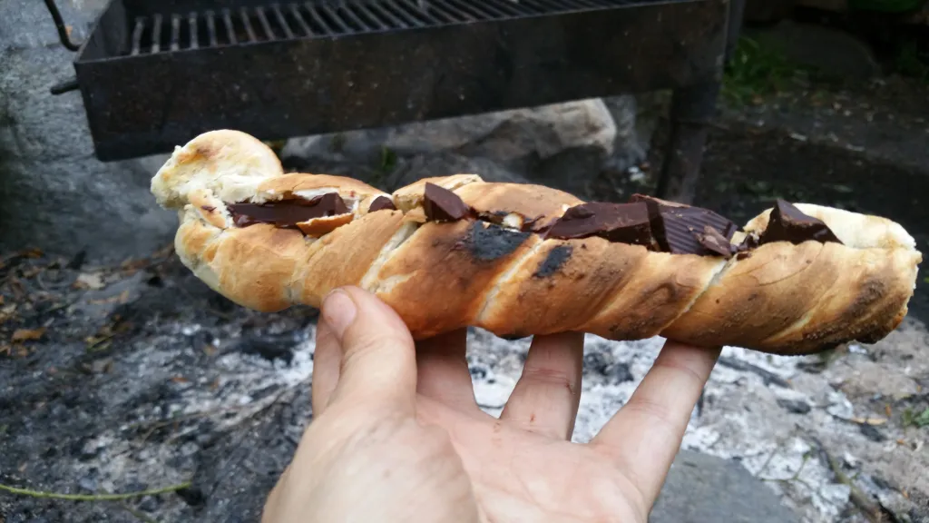 En hånd holder et snobrød med mørk chokolade foran resterne af et bål