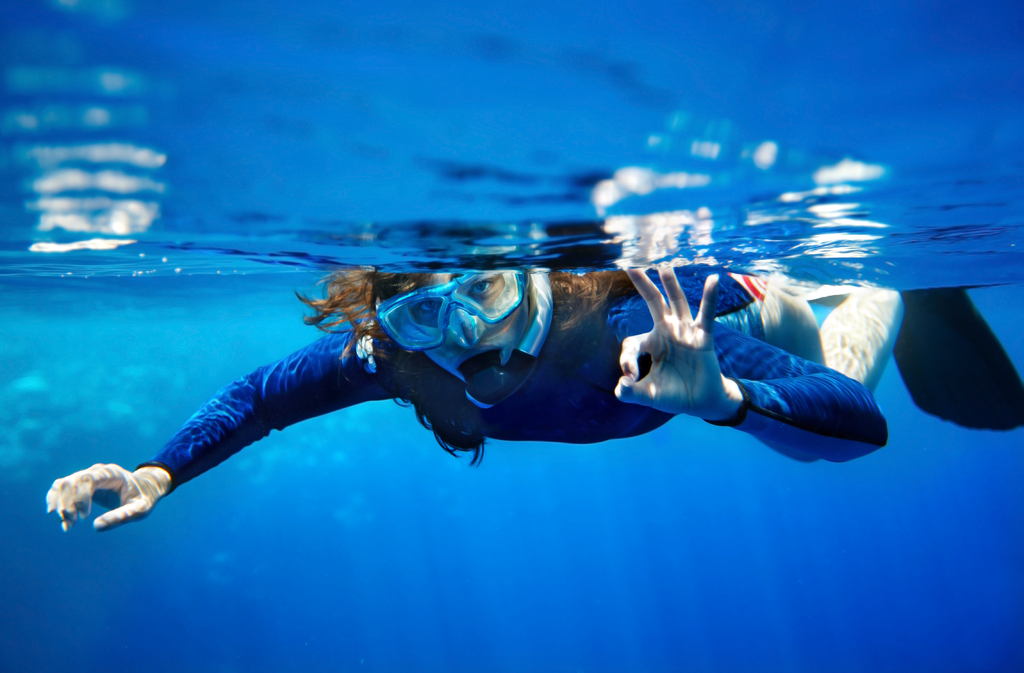 Kvinde med maske snorkler i klart, blåt vand og giver tegnet til at alt er godt